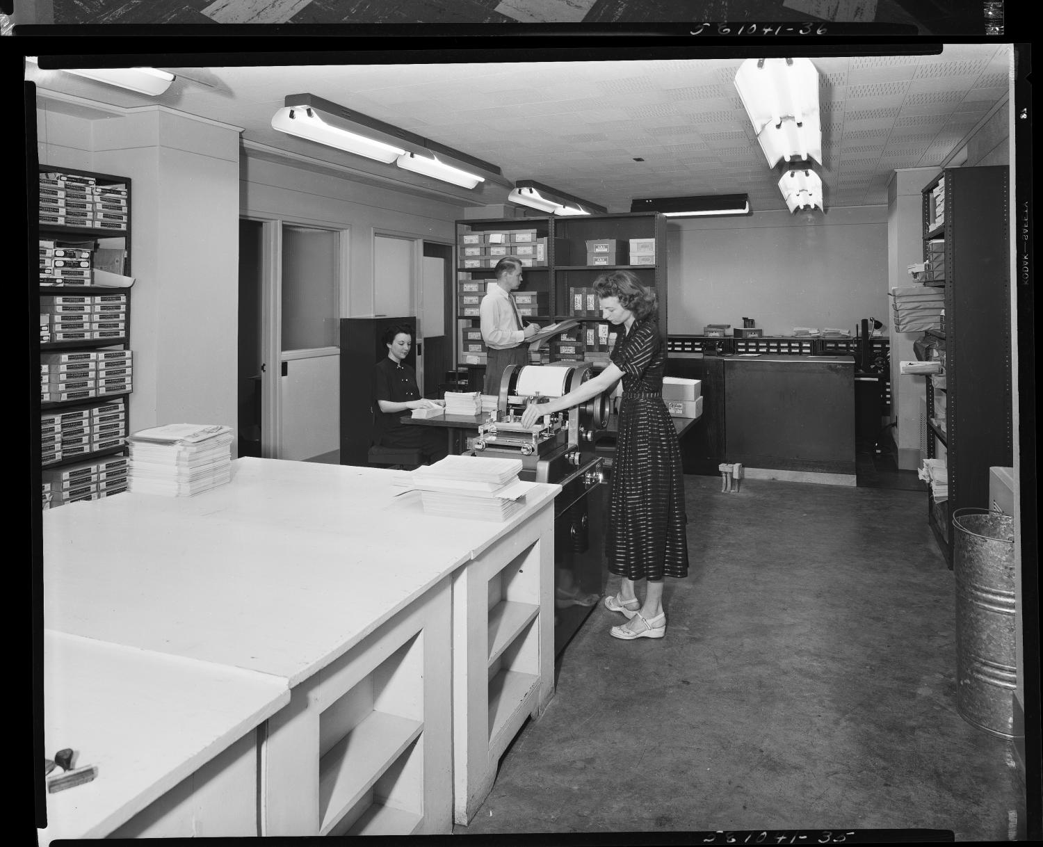 May 1950: the basement print shop, with a woman feeding a duplicating press, stacks of paper on a work table, and shelves of supplies